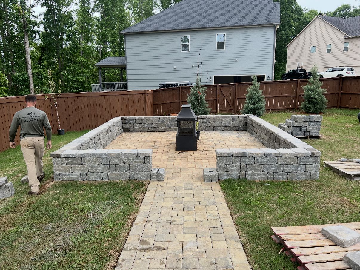 Paver patio enclosed by a gray block seat wall with a built-in fire feature and young evergreen plantings, connected by a paver walkway